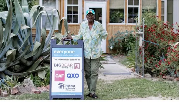 A homeowner standing beside a Habitat for Humanity project sign listing GAF as a project sponsor.