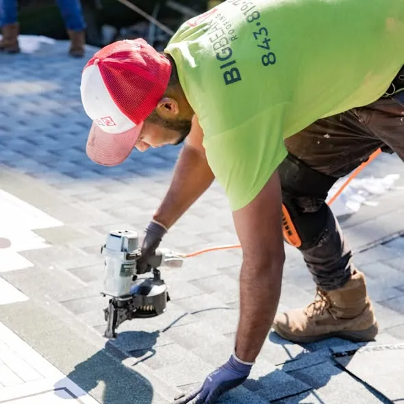 A roofer fastening shingles with a coil nailer on a sunny roof.